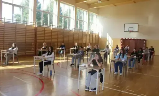 Młodzież siedzi w ławkach, pojedynczo, rozstawionych w odległościach na sali gimnastycznej, jak na maturach